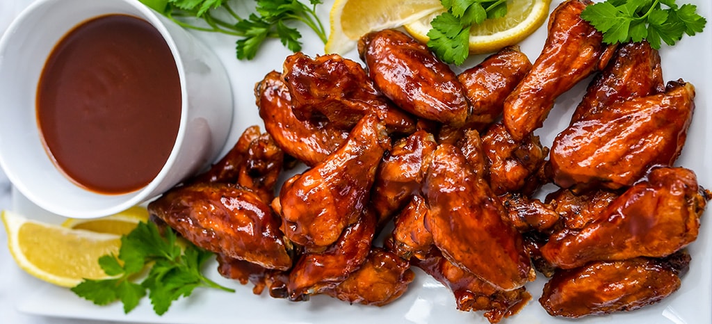 Delicious barbecue wings along with a cup of sauce, parsley, and lemon all served on a plate.
