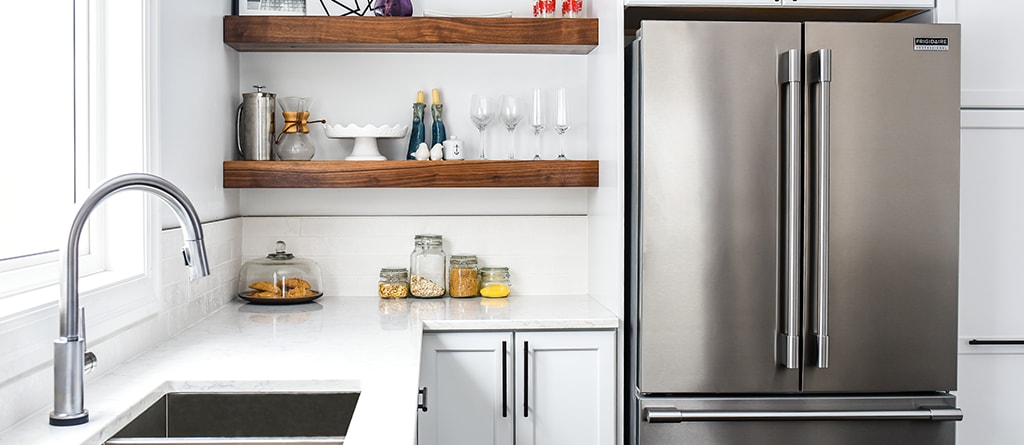 Kitchen setting with stocked natural wood shelves, white marble counter tops, and a stainless-steel refrigerator.