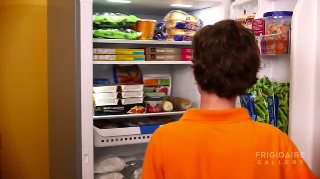 Boy in an orange shirt looking into an open and fully stocked refrigerator.