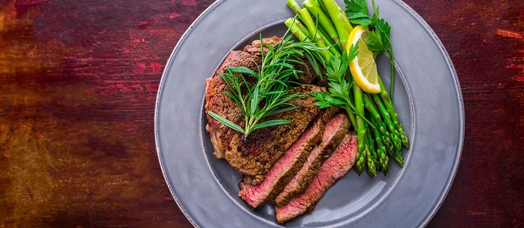 Thick medium-rare boneless rib eye steak under fresh rosemary and fresh garlic cloves with asparagus on a serving dish.  Recipe calls for 13 ingredients, 15 minutes cooking time, 20 minutes prep time, and 4 servings.