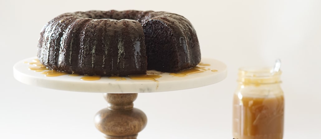 Rich and decadent Chocolate Bundt cake covered in a salted caramel sauce on a marble pedestal.  Recipe calls for 14 ingredients, 70 minutes cooking time, 20 minutes prep time, and 12 servings.