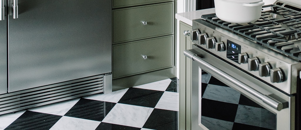 Stainless steel refrigerator next to a Gas stove in a kitchen with white and black tiled floor.